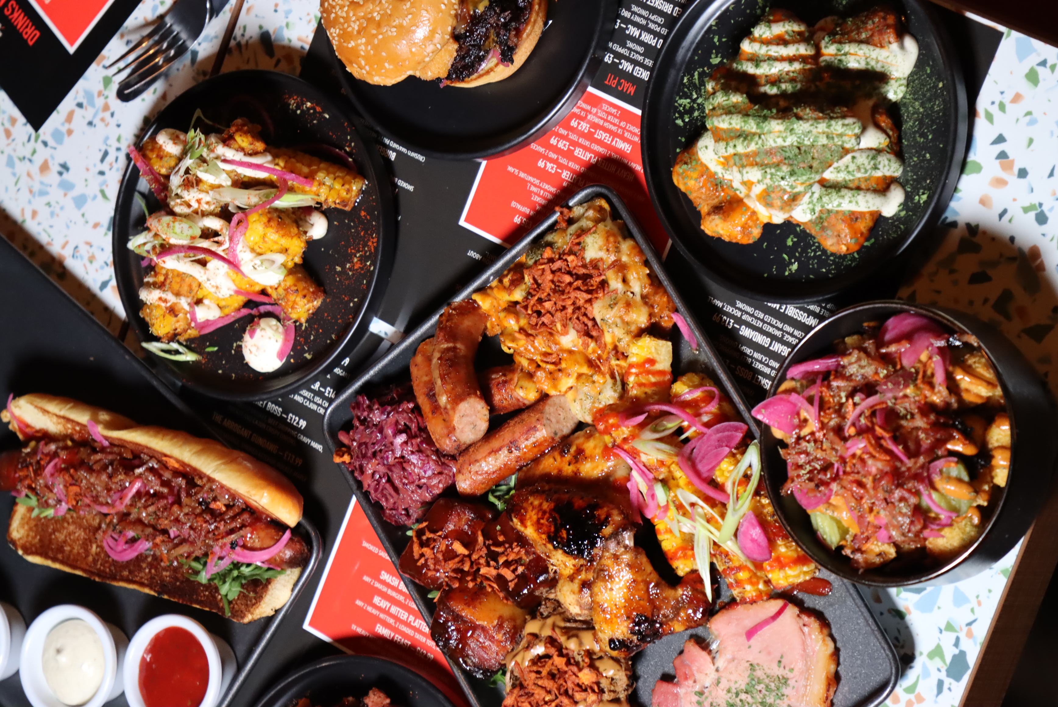Variety of grilled and sauced dishes including a loaded hot dog, corn on the cob with toppings, a burger, chicken wings with sauce, sausages, and poutine with pickled onions on black plates and trays.