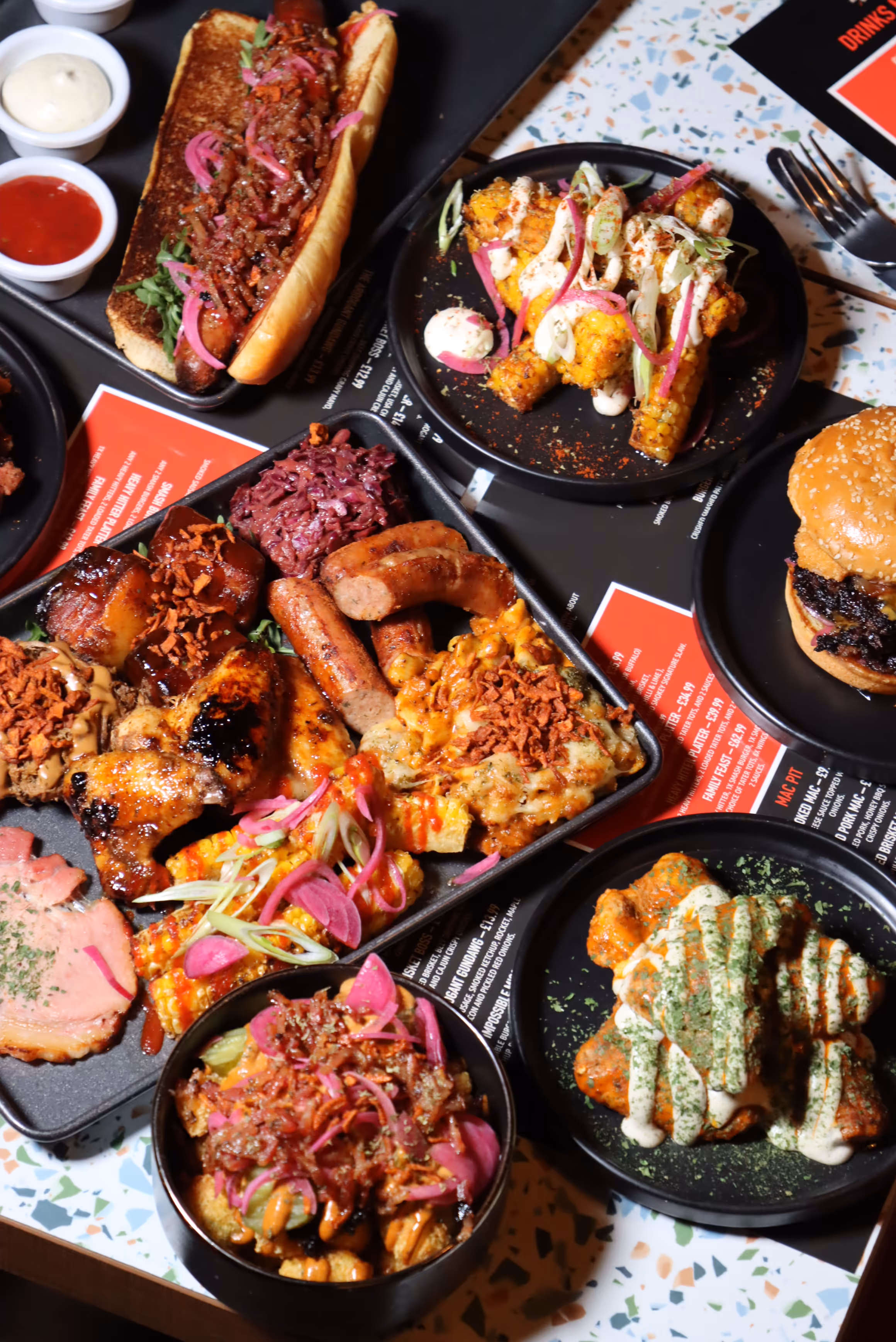Variety of grilled and sauced dishes including a loaded hot dog, corn on the cob with toppings, a burger, chicken wings with sauce, sausages, and poutine with pickled onions on black plates and trays.