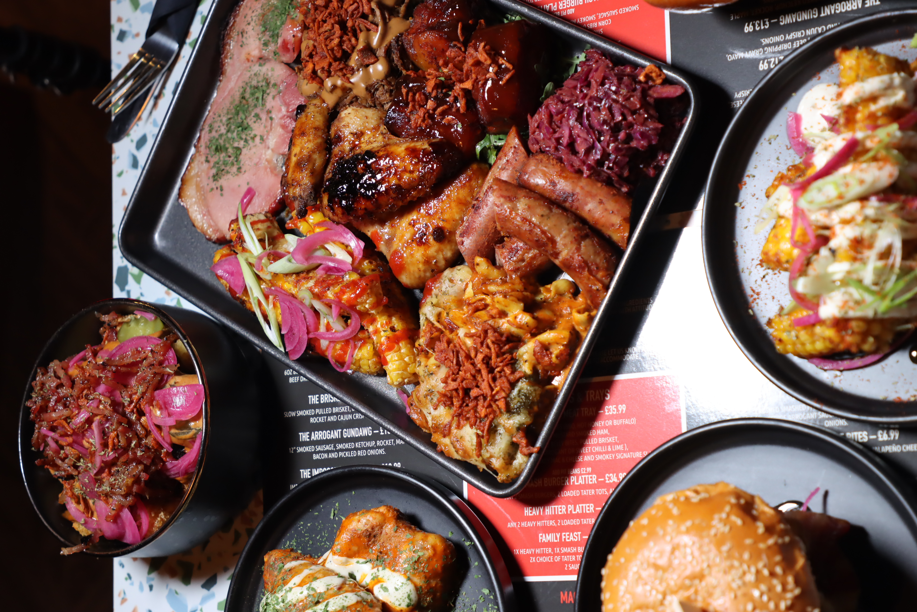 Tray of assorted grilled meats, sausages, mac and cheese, corn on the cob, pickled red onions, and coleslaw with surrounding plates of a burger, sauced wings, and loaded fries topped with pickled onions and bacon bits.