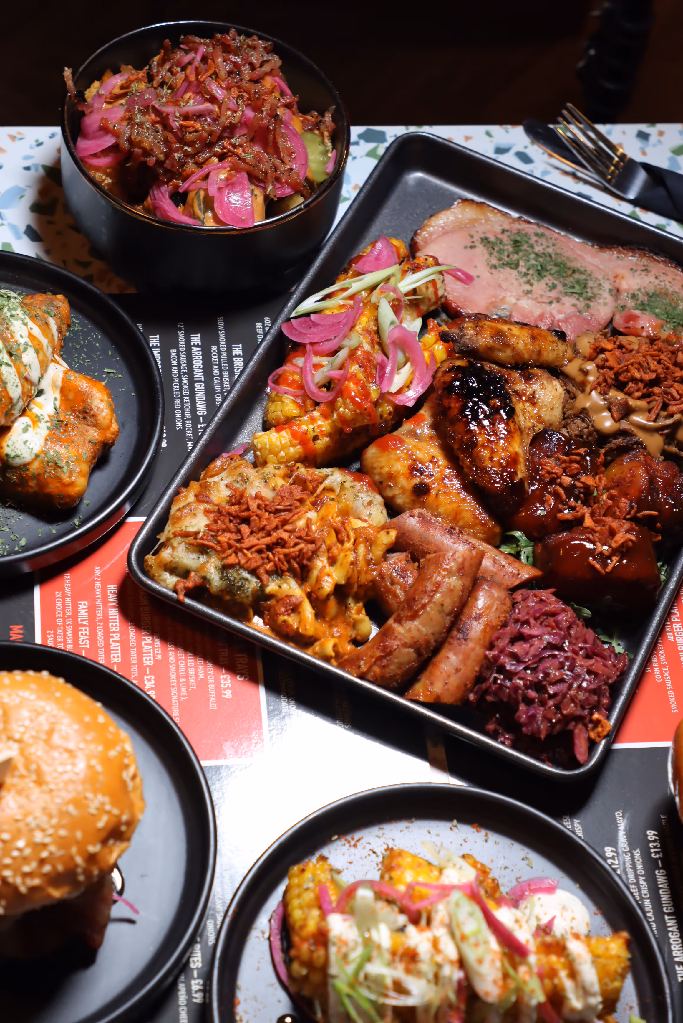 Tray of assorted grilled meats, sausages, mac and cheese, corn on the cob, pickled red onions, and coleslaw with surrounding plates of a burger, sauced wings, and loaded fries topped with pickled onions and bacon bits.