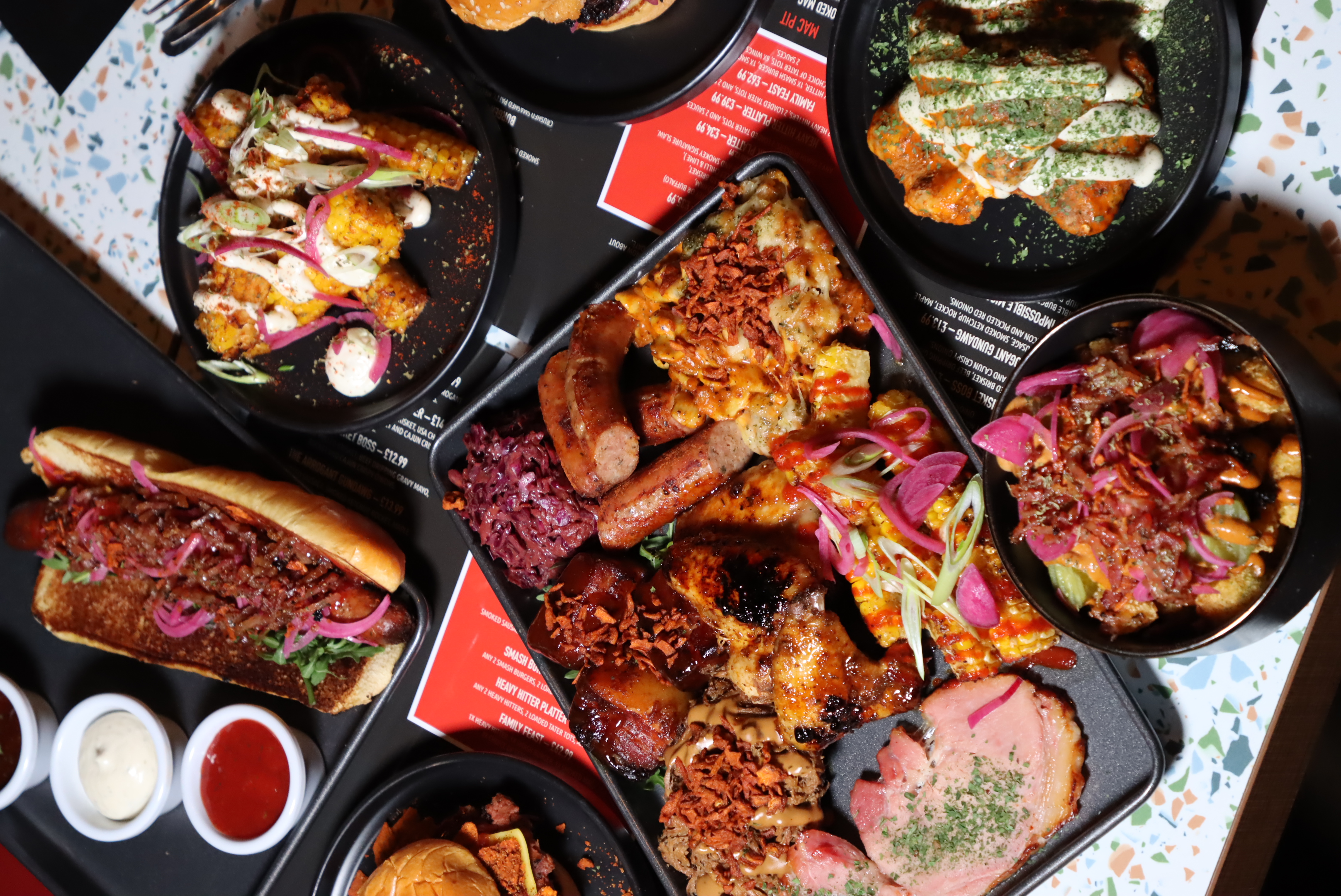 Assortment of grilled barbecue dishes including sausages, chicken wings, pulled pork, cornbread with toppings, coleslaw, a hot dog with relish, and various sauces on black plates.