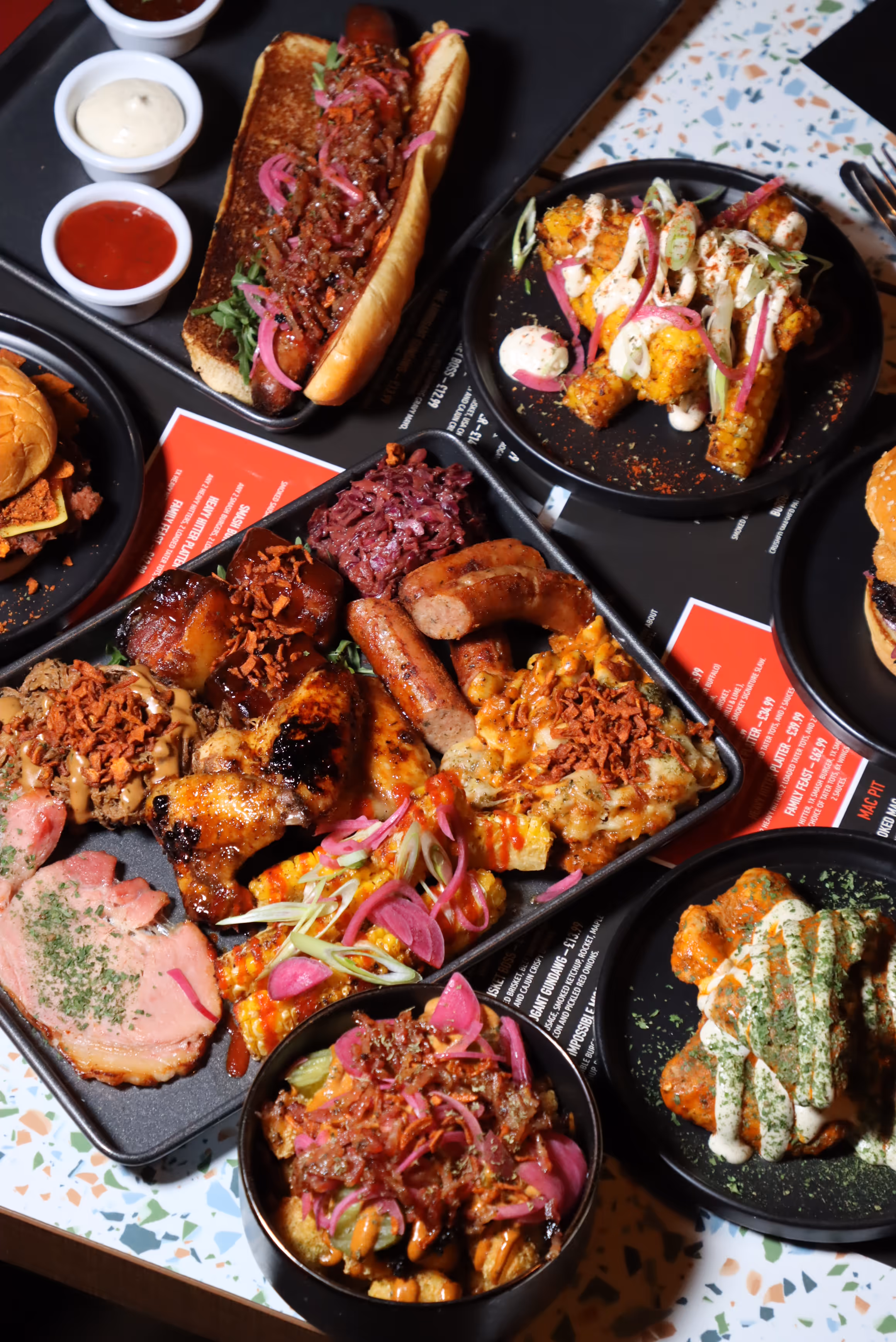 Assortment of grilled barbecue dishes including sausages, chicken wings, pulled pork, cornbread with toppings, coleslaw, a hot dog with relish, and various sauces on black plates.