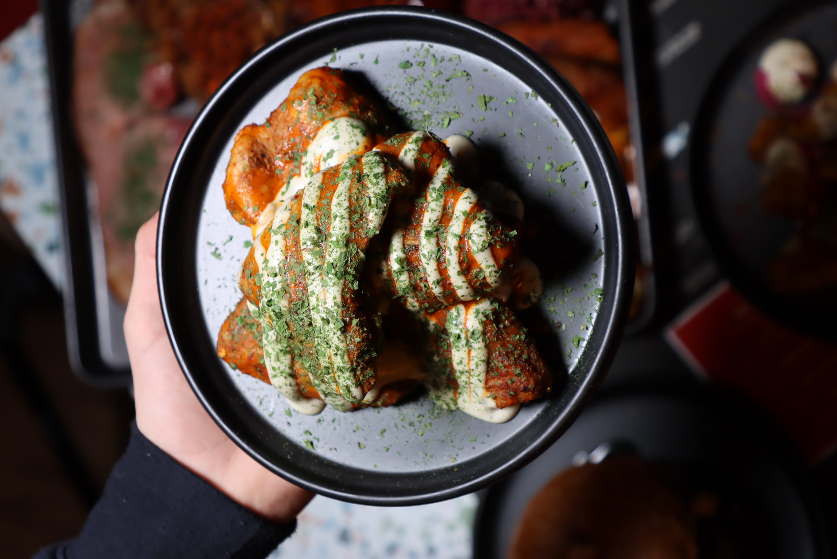 Hand holding a black plate with four pieces of crispy fried chicken drizzled with creamy sauce and sprinkled with chopped herbs.