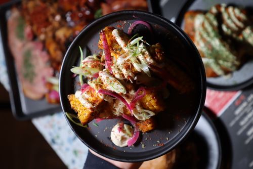 Grilled corn on the cob pieces topped with creamy sauce, sliced red onions, and green herbs in a black bowl.
