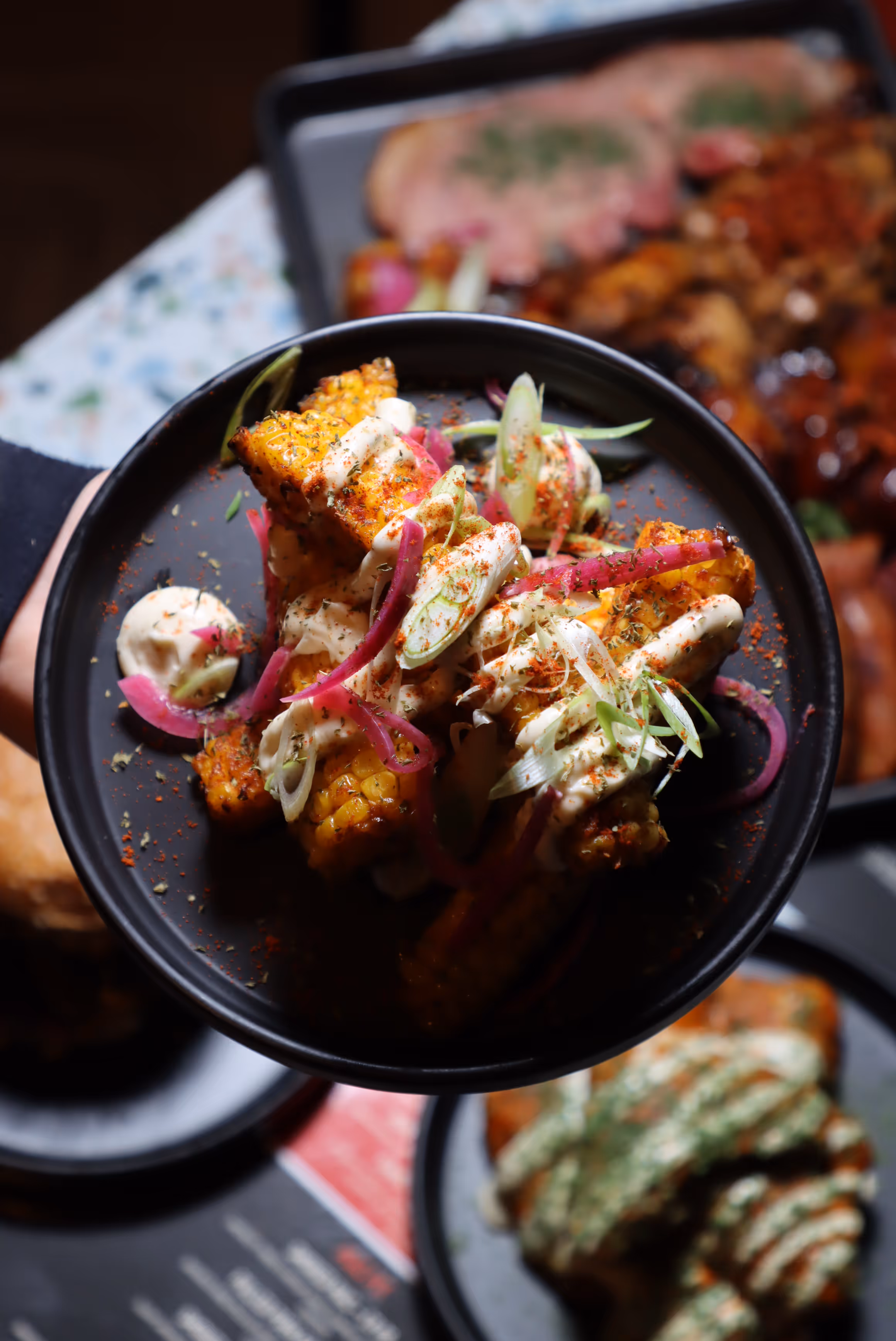 Grilled corn on the cob pieces topped with creamy sauce, sliced red onions, and green herbs in a black bowl.