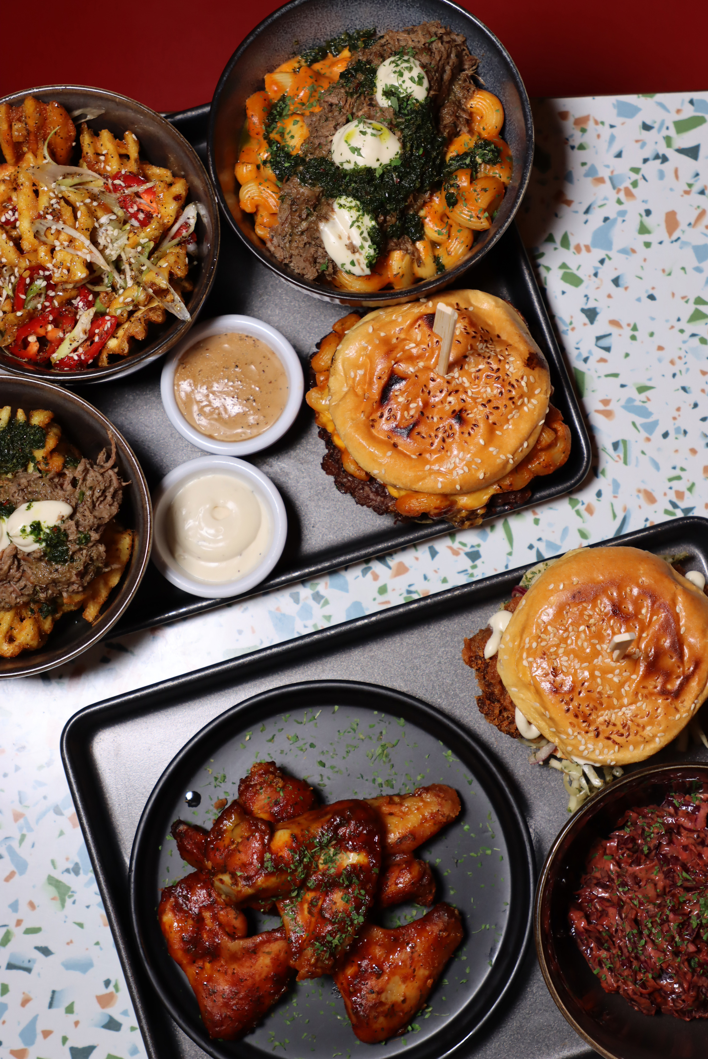 Tray with two burgers, bowls of loaded waffle fries topped with herbs and sauces, and a plate of glazed chicken wings garnished with parsley.
