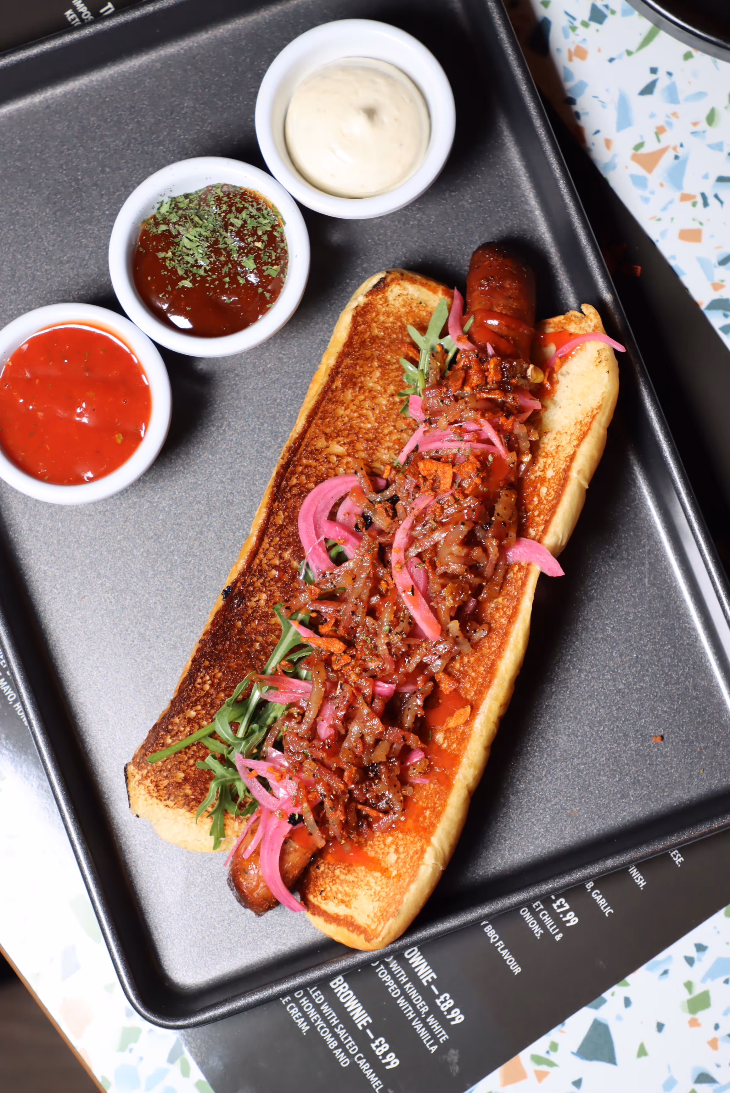 Grilled hot dog bun with a sausage topped with pickled red onions, crispy bacon bits, and greens, served on a tray with three small bowls of different dipping sauces.