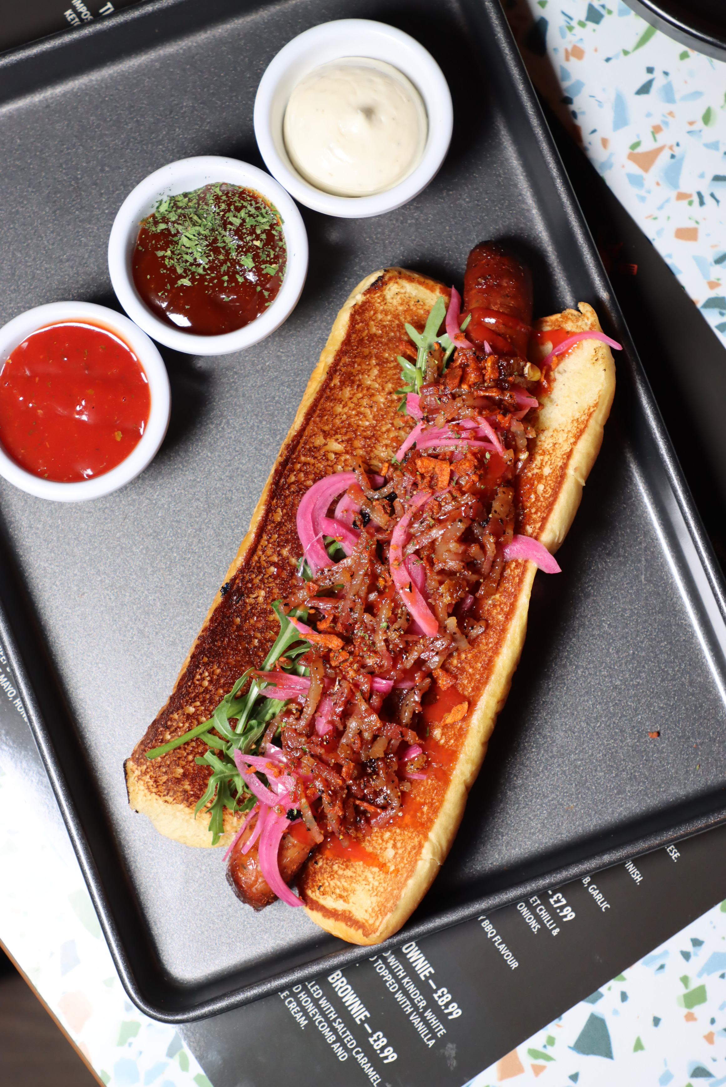 Grilled hot dog bun with a sausage topped with pickled red onions, crispy bacon bits, and greens, served on a tray with three small bowls of different dipping sauces.