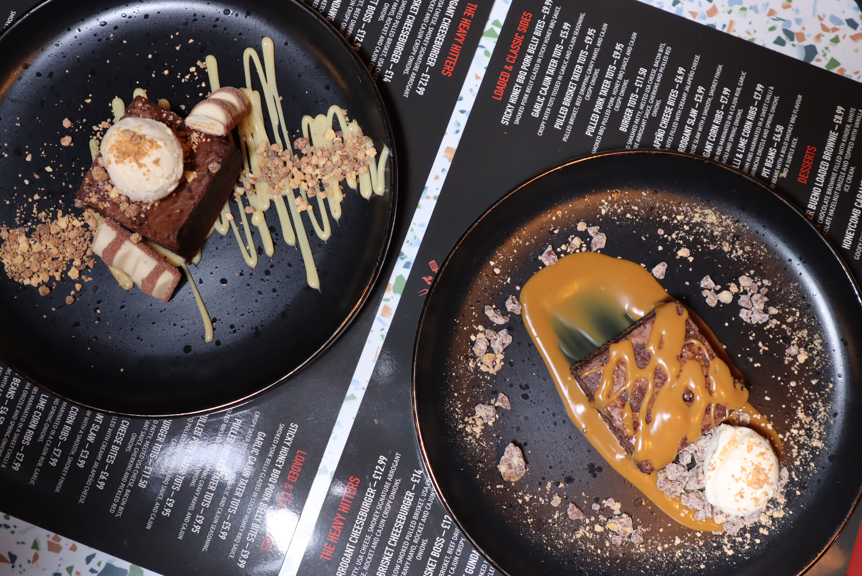 Two plates with brownies drizzled with caramel and cream sauce, garnished with crushed nuts and a scoop of vanilla ice cream on each, placed on a restaurant menu.