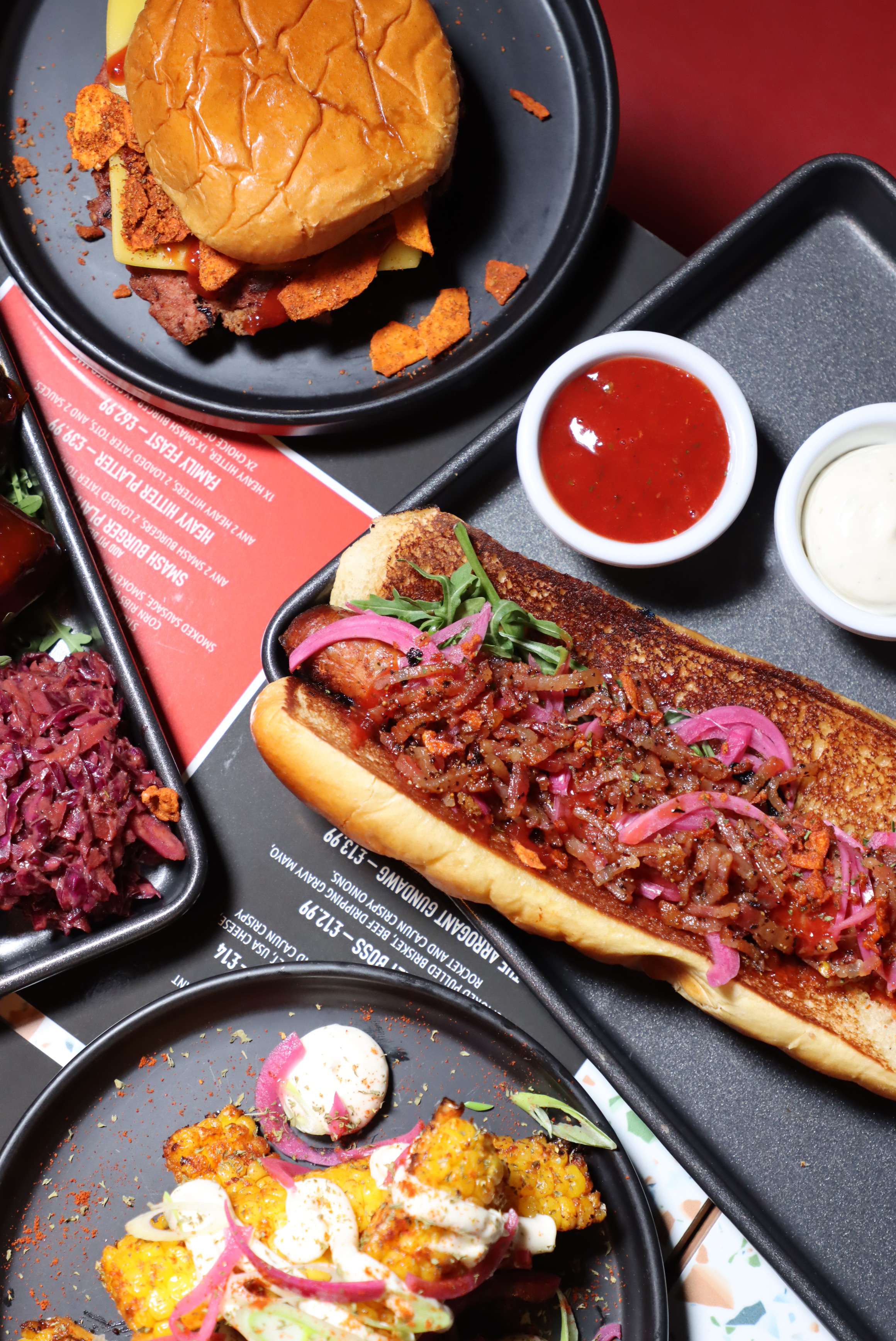 Assorted gourmet dishes including a burger with cheese and chips, a hot dog with onions and greens, grilled corn with sauce, and a side of red cabbage slaw on black plates.