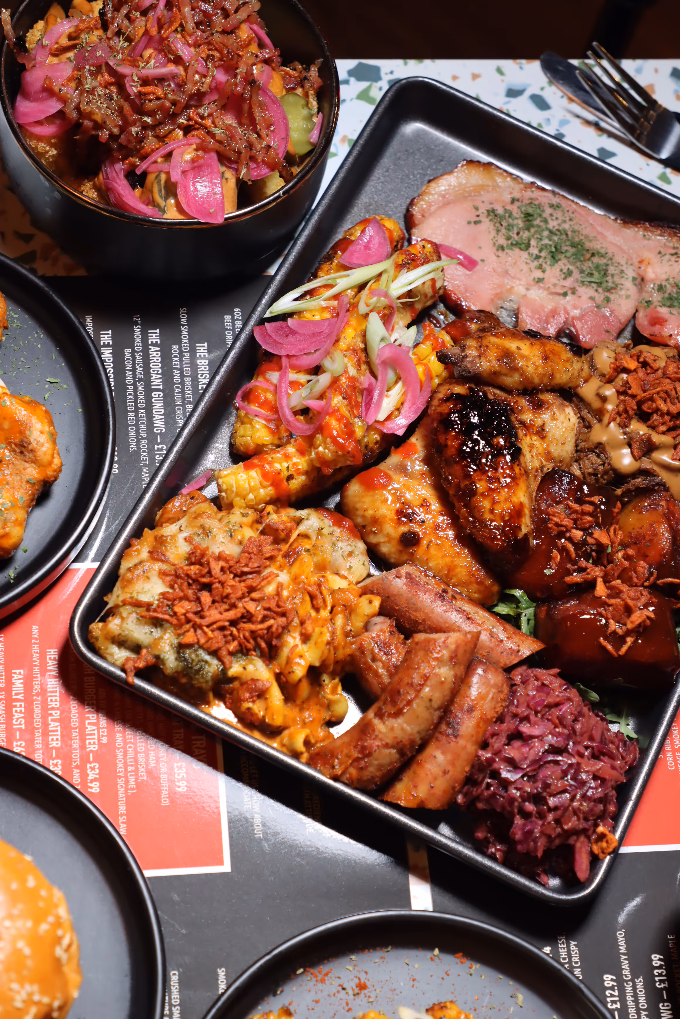 Tray of smoked barbecue platter with corn on the cob, smoked chicken wings, sausages, coleslaw, pork ribs, and macaroni and cheese topped with crispy onions and pickled red onions.
