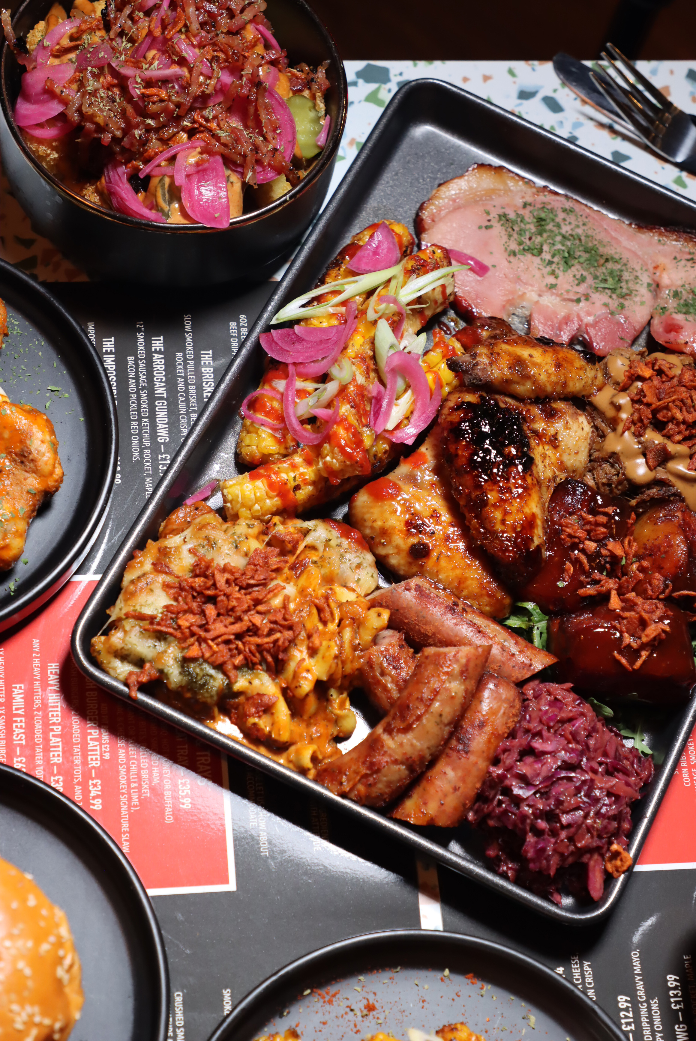 Tray of smoked barbecue platter with corn on the cob, smoked chicken wings, sausages, coleslaw, pork ribs, and macaroni and cheese topped with crispy onions and pickled red onions.