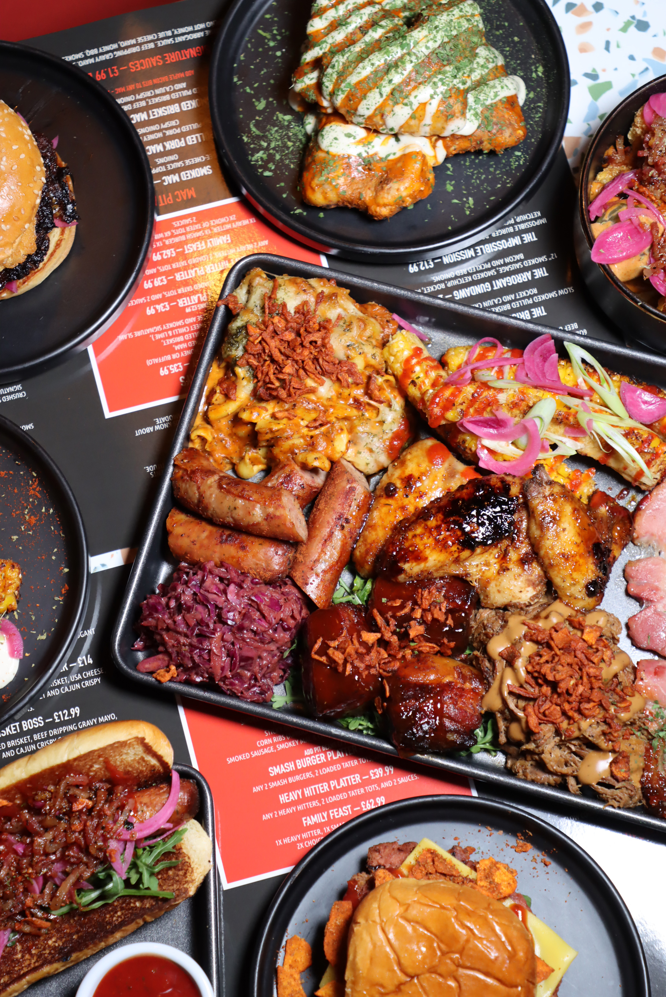 Variety of grilled and smoked meats including sausages, ribs, chicken wings, and burgers served on black plates and trays with garnishes and sauces.