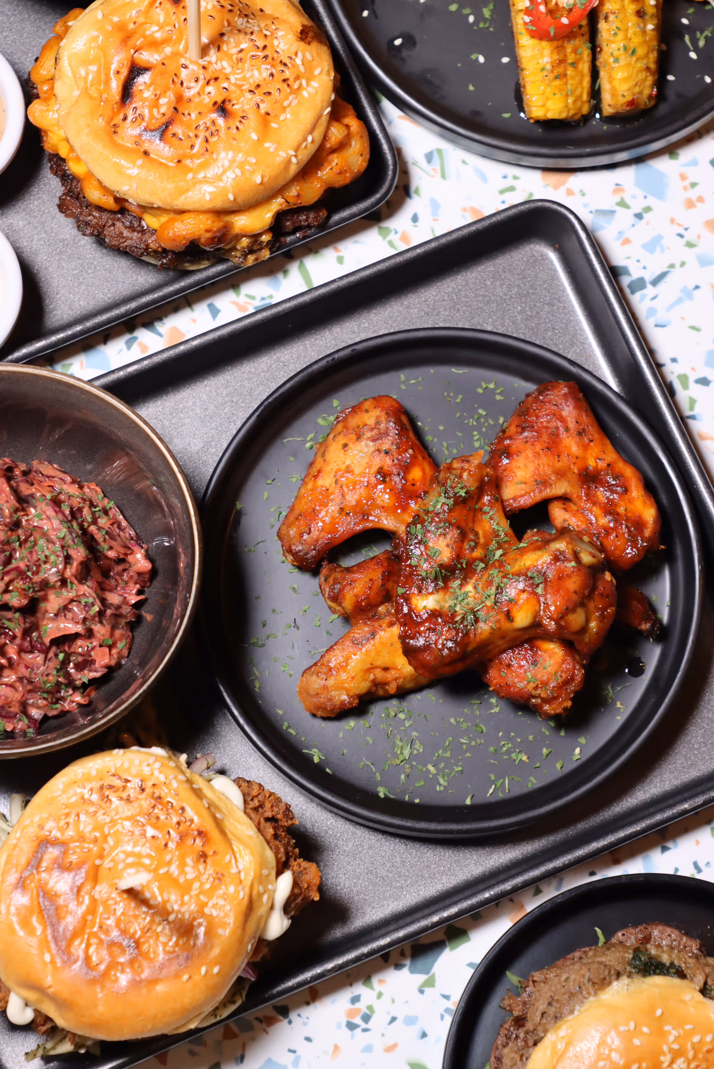 Black plates and trays with grilled chicken wings sprinkled with herbs, burgers with sesame seed buns, corn on the cob, and a bowl of coleslaw, arranged on a terrazzo surface.
