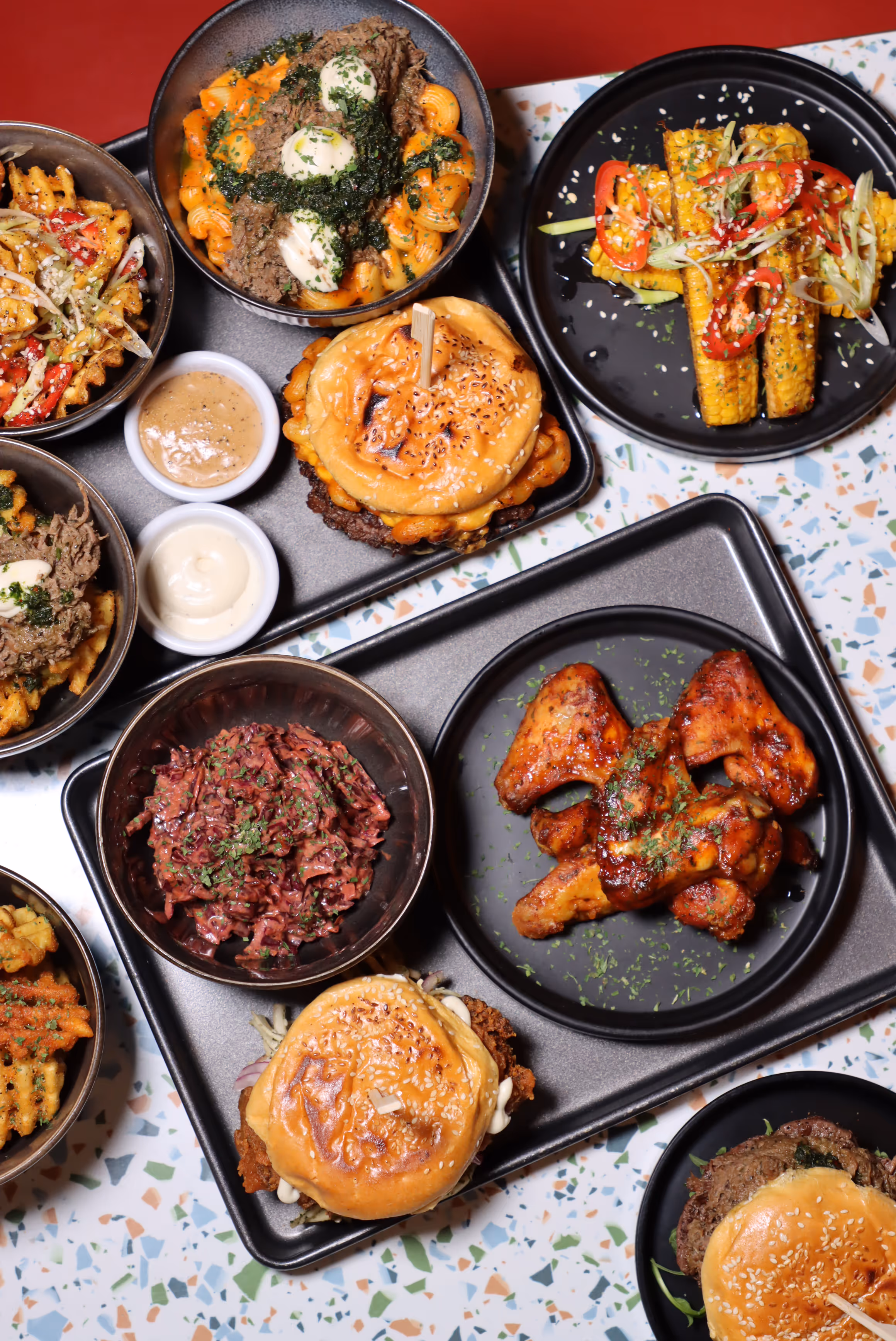 Overhead view of a variety of plated foods including two burgers, spicy chicken wings, waffle fries with sauces, grilled corn with sesame seeds and red peppers, and bowls of shredded beef and coleslaw on a terrazzo tabletop.