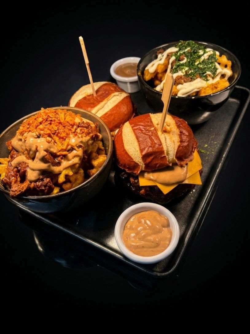 Tray with two cheeseburgers on pretzel buns, two bowls of waffle fries topped with cheese sauce and toppings, and two small containers of dipping sauce.