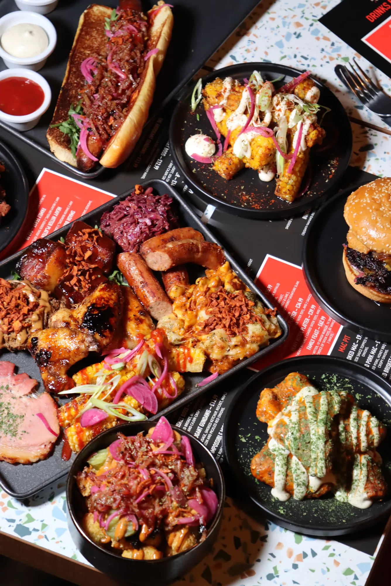 Variety of grilled and sauced dishes including a loaded hot dog, corn on the cob with toppings, a burger, chicken wings with sauce, sausages, and poutine with pickled onions on black plates and trays.