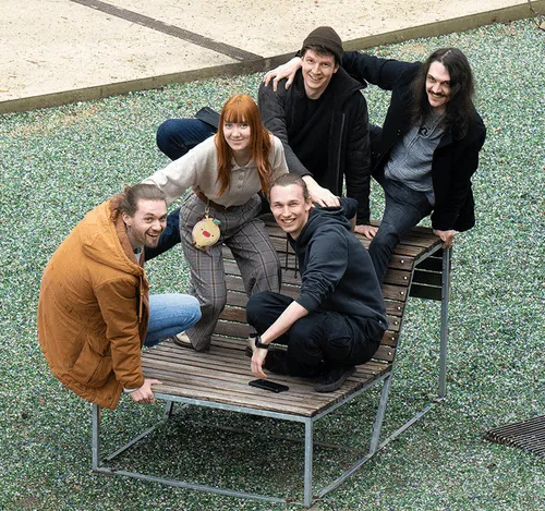 Group of five young adults smiling and sitting closely together on and around a unique metal and wooden bench outdoors.