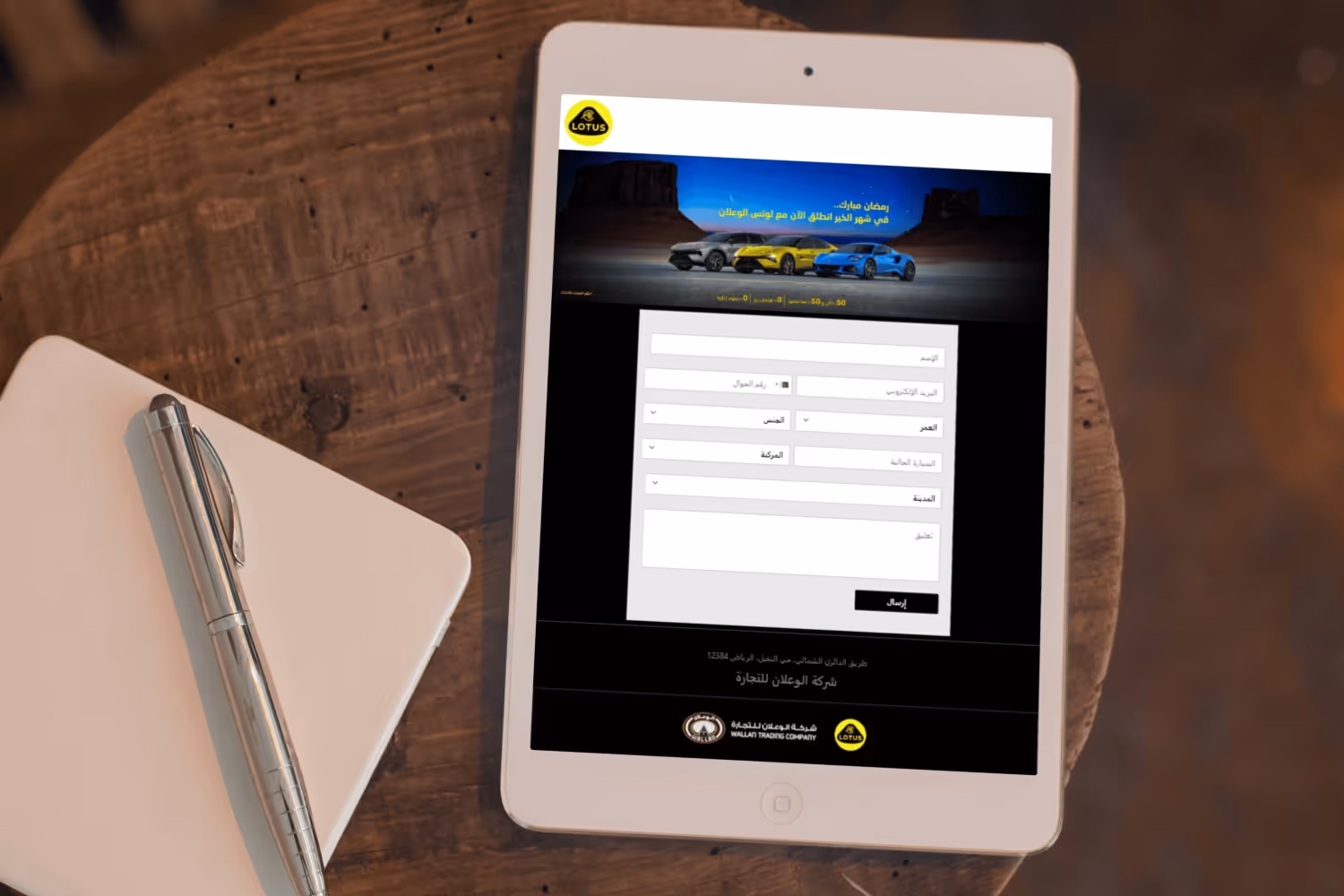 Tablet on wooden table displaying a Lotus promotional form in Arabic with images of three cars and input fields for name, email, age, gender, car model, city, and comment.