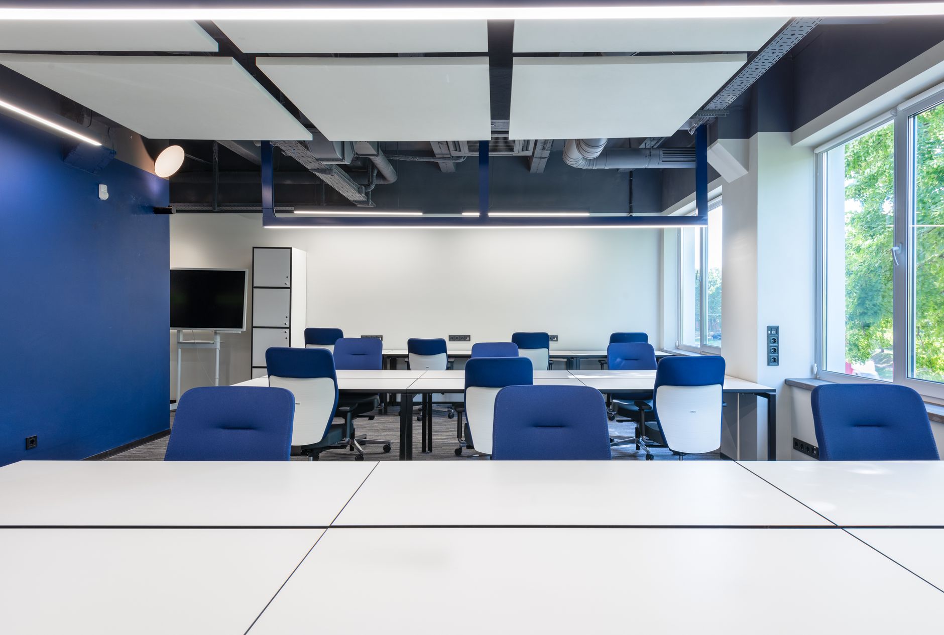 Various white desks with blue chairs placed in modern spacious workspace with big window and monitor in contemporary business