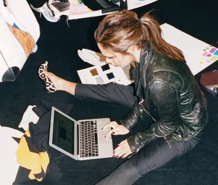 A woman sits on the floor working on a laptop, surrounded by design materials and swatches, wearing a leather jacket and stylish shoes.