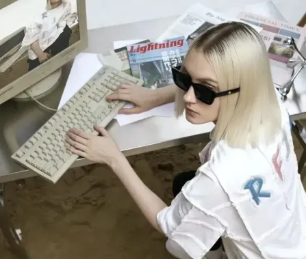 A person with blonde, straight hair sits at a computer, wearing sunglasses and typing on an old keyboard, surrounded by magazines.