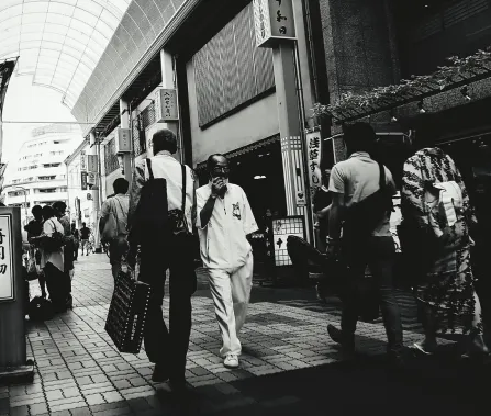 A busy street scene in black and white, featuring various pedestrians walking along a covered walkway lined with shops.