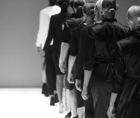 Fashion models walk down a runway in a line, showcasing elegant black and white outfits against a minimalist backdrop.