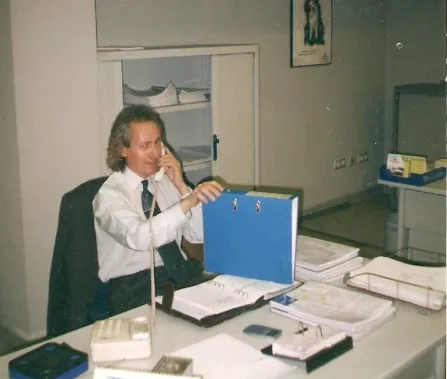 A man in a suit sits at a desk, talking on the phone, with folders and documents arranged around him in an office setting.