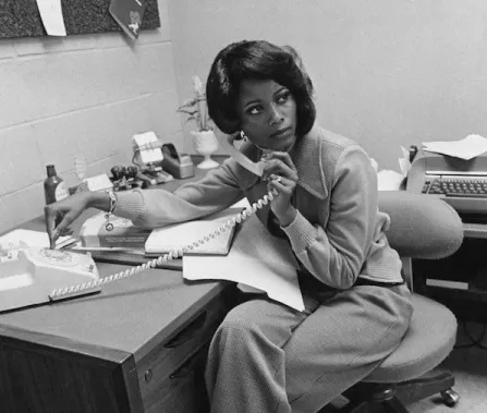 Black and white image of a woman seated at a desk, talking on a rotary phone, surrounded by paperwork and a typewriter.