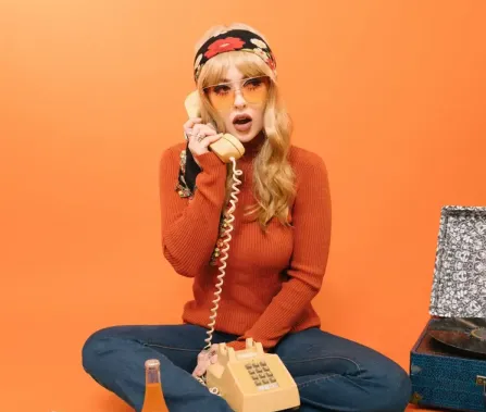 A woman in a bright orange sweater with a floral headband sits on the floor talking on a vintage telephone against an orange background.