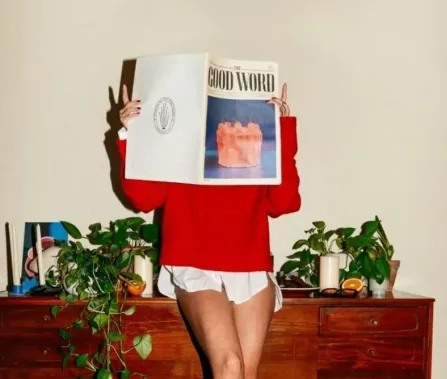 A person in a red sweater holds a magazine titled "THE GOOD WORD" in front of their face, standing by a wooden dresser with plants.