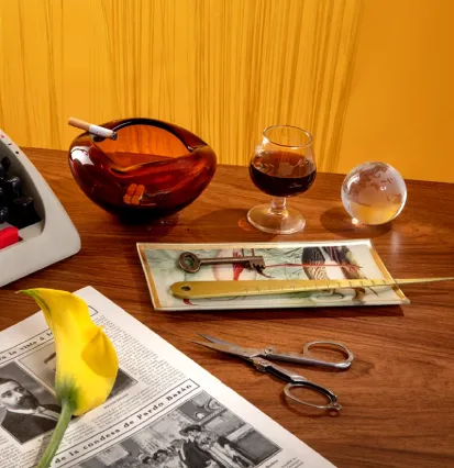 A vintage desk scene featuring a typewriter, an amber ashtray, a glass, a crystal ball, scissors, a newspaper, and a yellow flower.