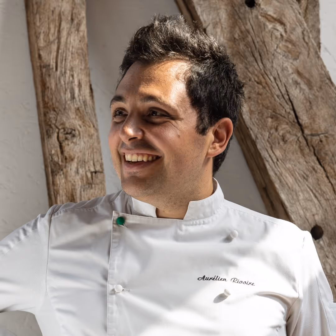 Portrait d'un homme souriant portant une veste de chef blanc avec le nom Aurelien Rivoire brodé, devant un mur avec des poutres en bois.