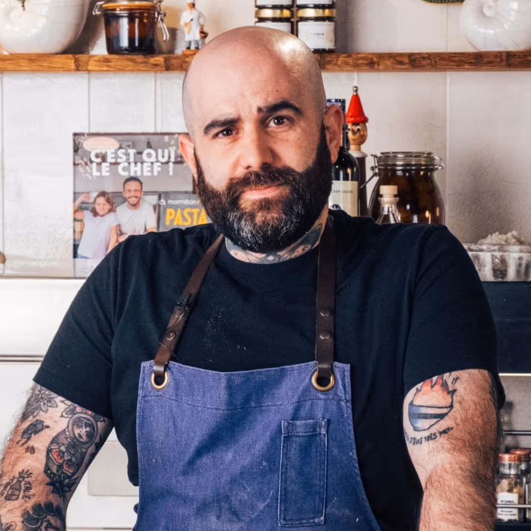 Chef barbu et chauve portant un tablier bleu dans une cuisine avec des tatouages visibles sur ses bras.