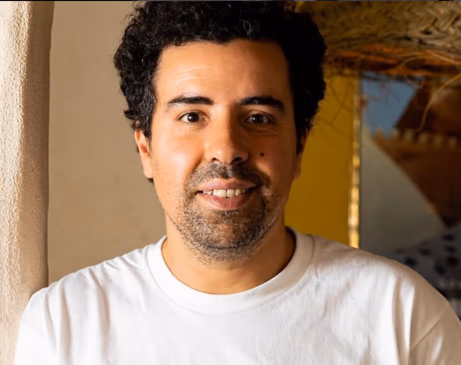 Homme souriant aux cheveux bouclés noirs et barbe légère portant un t-shirt blanc, devant un mur clair et une décoration floue.