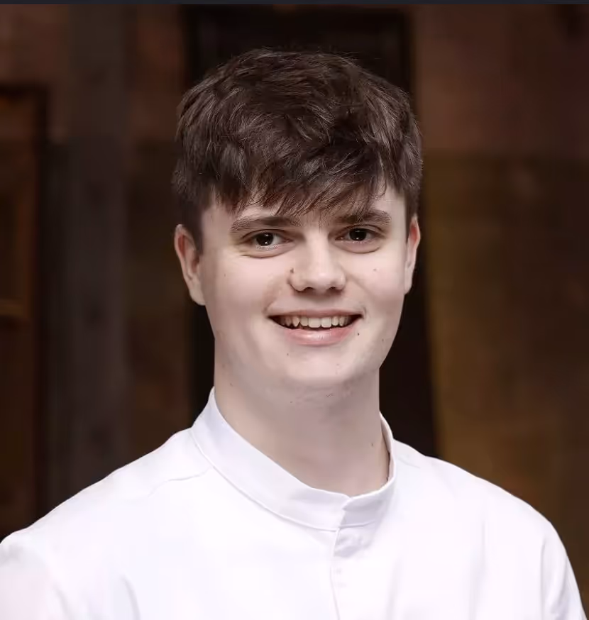 Jeune homme souriant avec une chemise blanche devant un fond flou sombre.