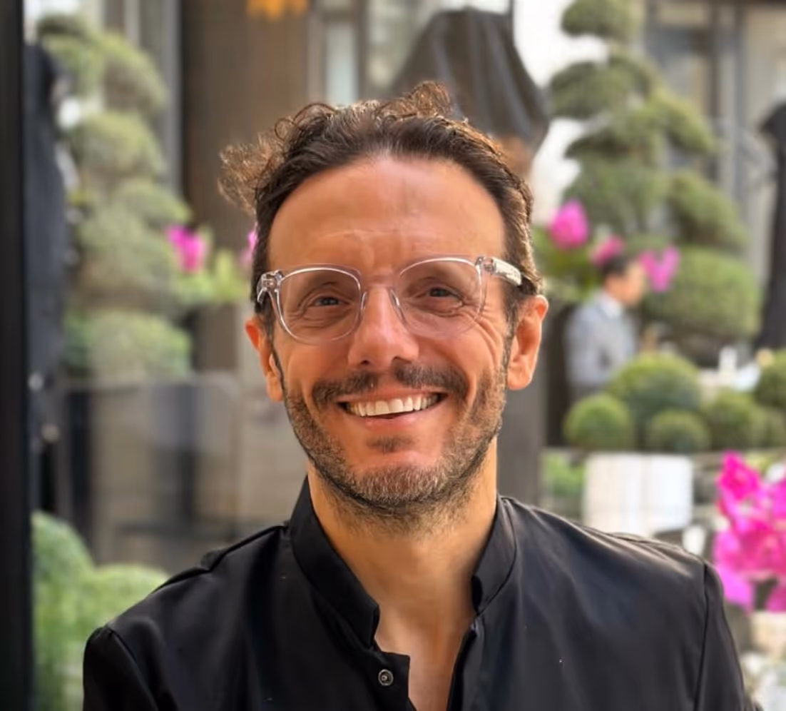 Homme souriant portant des lunettes transparentes et une veste noire, avec un fond flou de plantes et fleurs.
