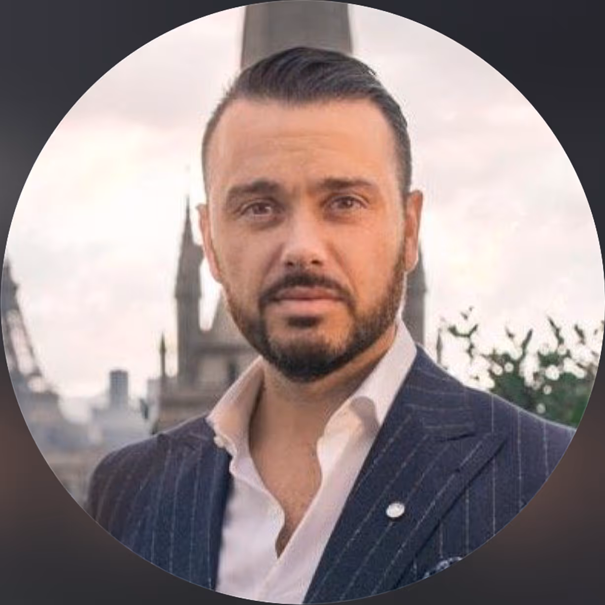 Homme aux cheveux foncés coiffés en arrière et barbe, portant un costume rayé bleu marine avec une chemise blanche, avec en arrière-plan un monument flou et un ciel nuageux.