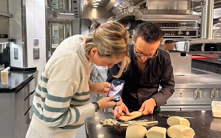 Une femme prend en photo un homme qui explique une technique avec de la pâte, dans une cuisine professionnelle.