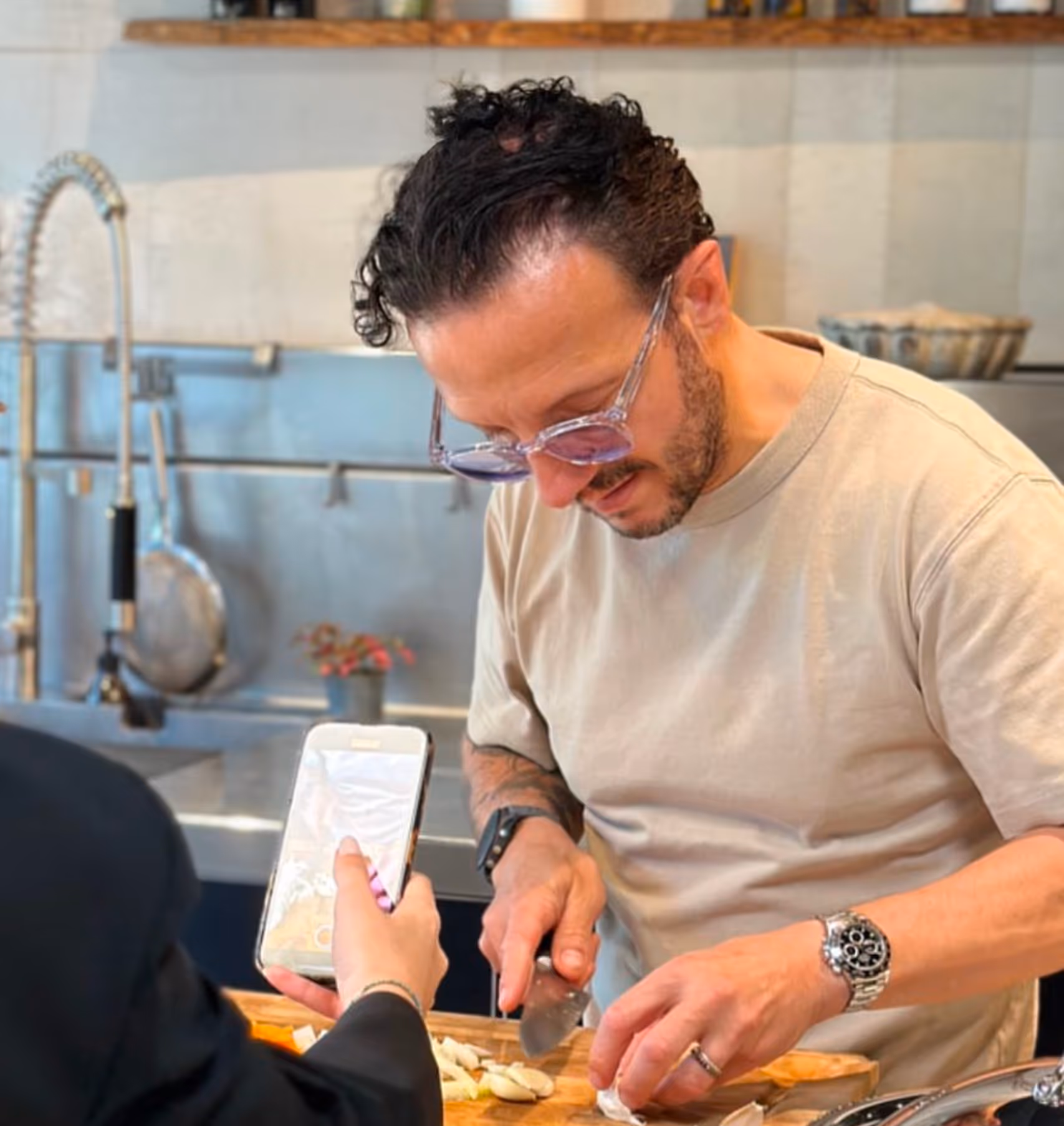 Un homme portant des lunettes coupe des oignons sur une planche à découper dans une cuisine, pendant qu'une autre personne tient un smartphone vers lui.