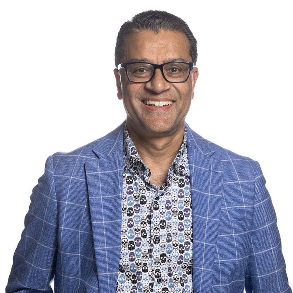 Smiling man wearing black glasses, a blue plaid blazer, and a skull-patterned shirt.