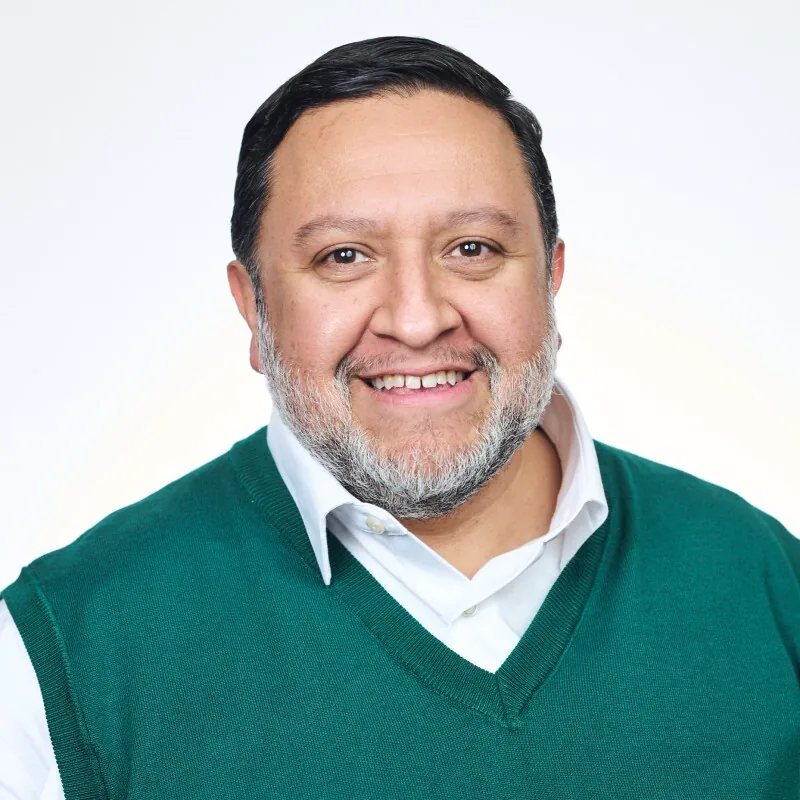 Smiling middle-aged man with short dark hair and salt-and-pepper beard wearing a green sweater vest over a white collared shirt.