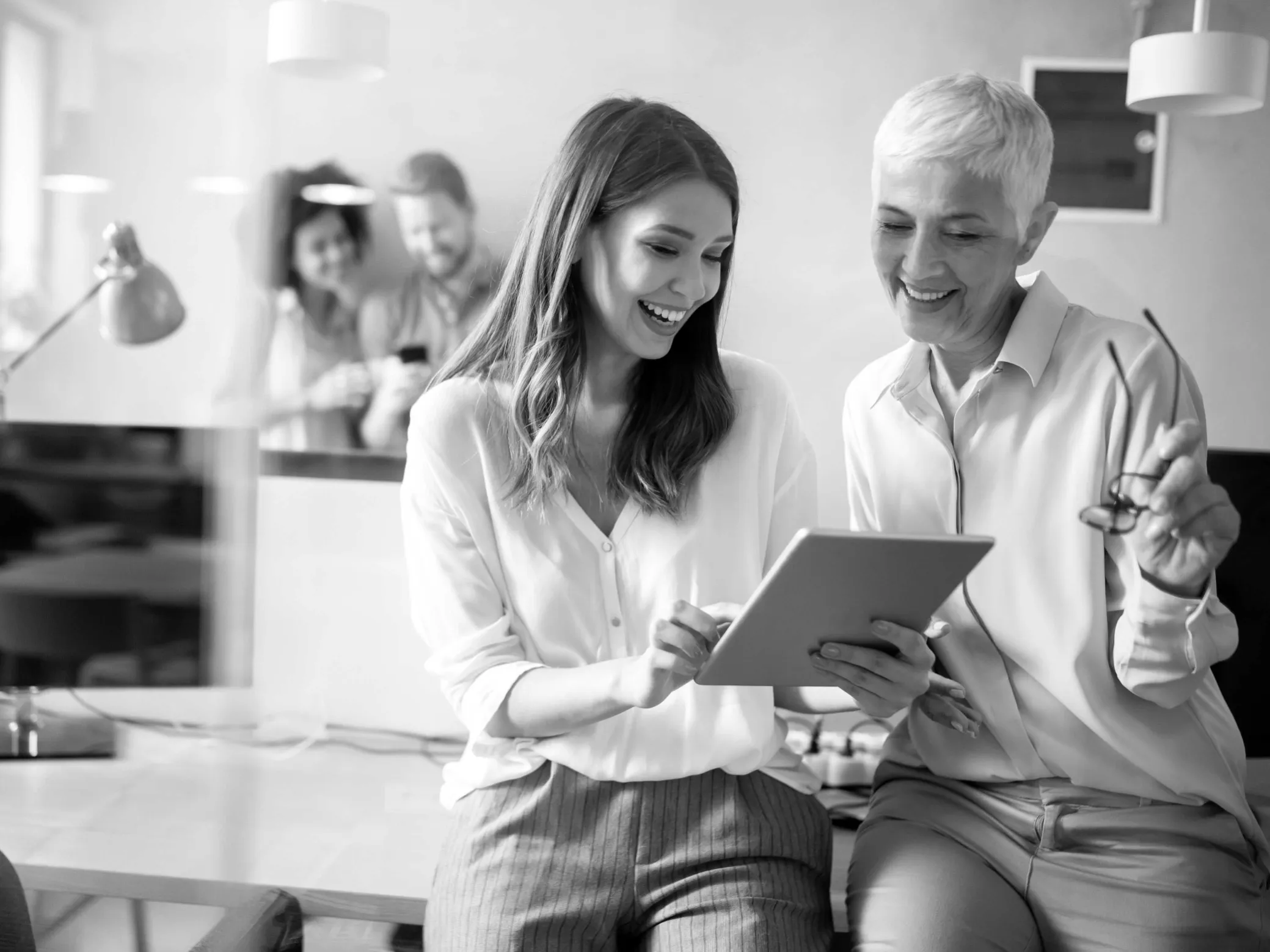 Two women sitting side by side smiling and looking at a tablet device, with a man and another woman blurred in the background.