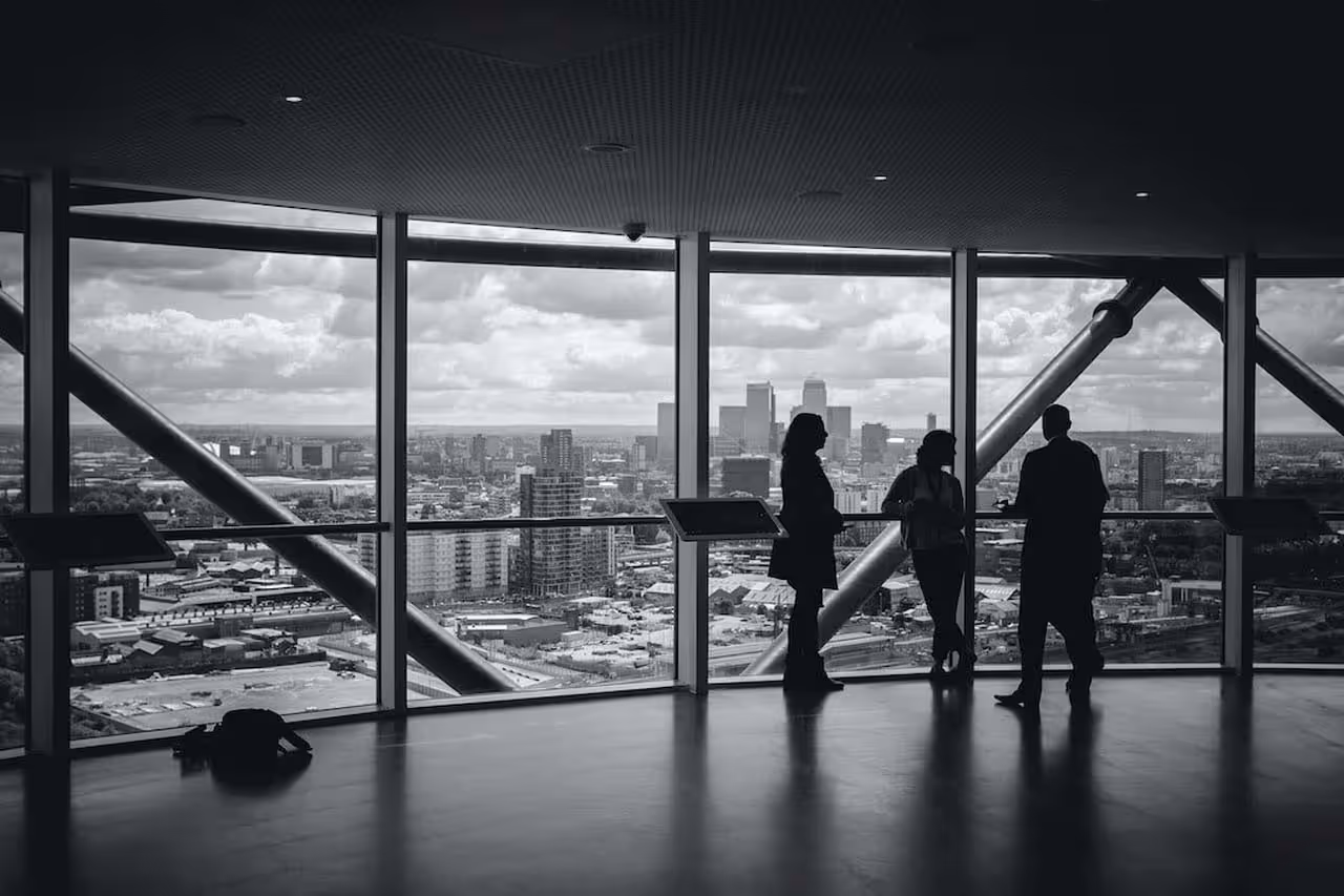 Silhouettes of three people talking inside a modern building with large windows overlooking a cityscape under a cloudy sky.