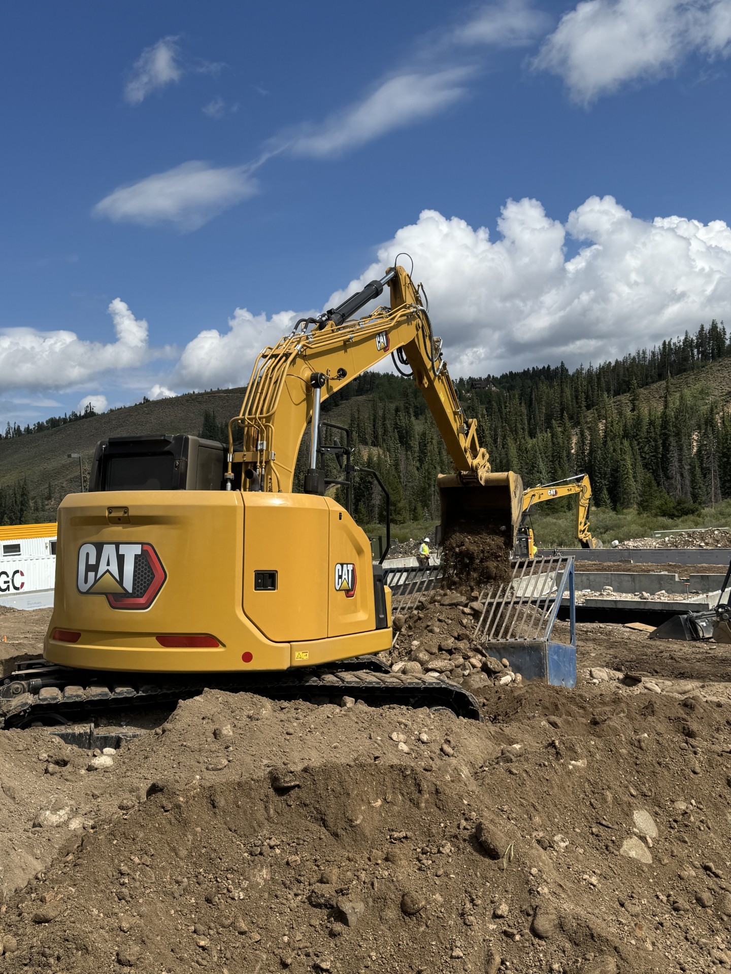 Yellow CAT excavator dumping dirt and rocks into a blue grading rake at a construction site with forested hills and partly cloudy sky in the background.