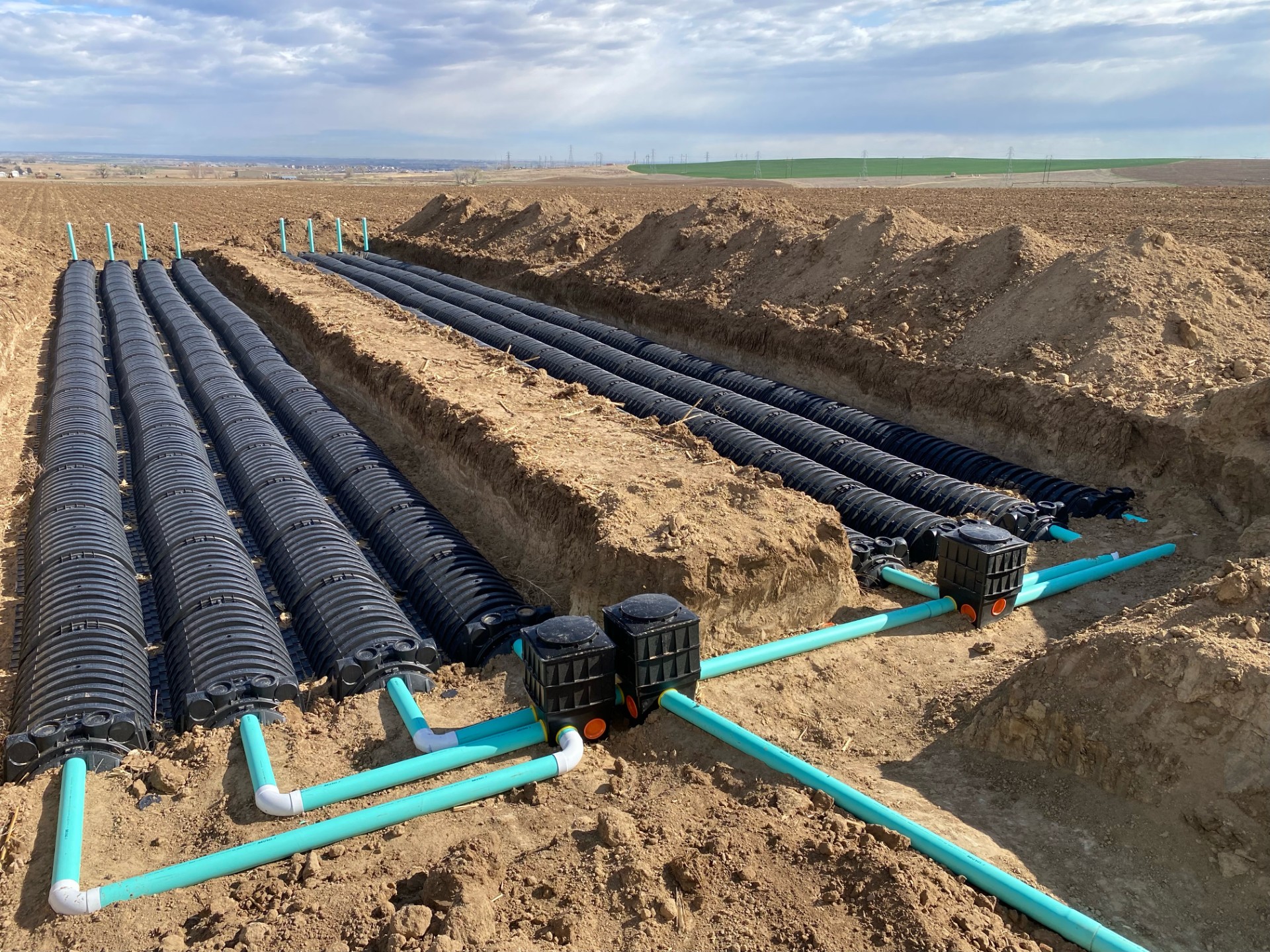 Underground drainage system installation with black corrugated pipes connected to blue PVC pipes in a large open field.