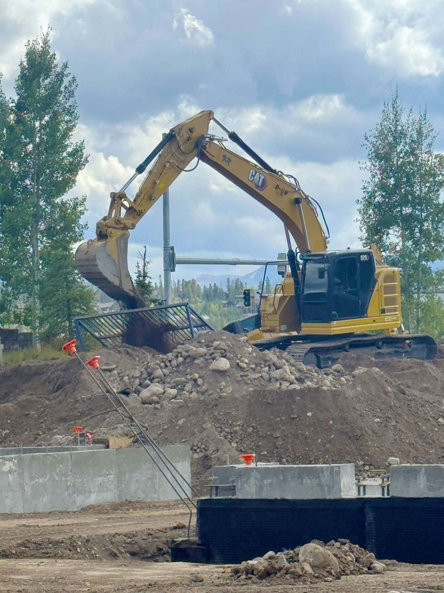 Yellow CAT excavator loading dirt and rocks onto a construction site with trees and cloudy sky in the background.