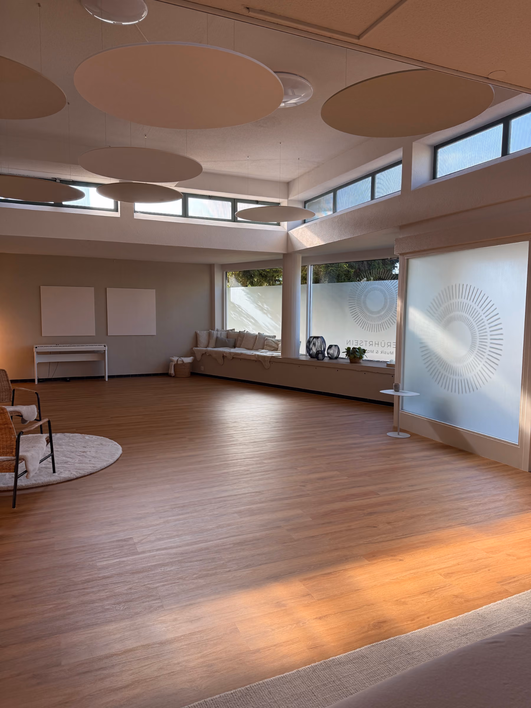 Der Raum Berührtsein. Ein großer heller Raum mit Holzboden, Sitzbank mit Kissen am Fenster und runden, an der Decke hängenden Akustikpaneelen.