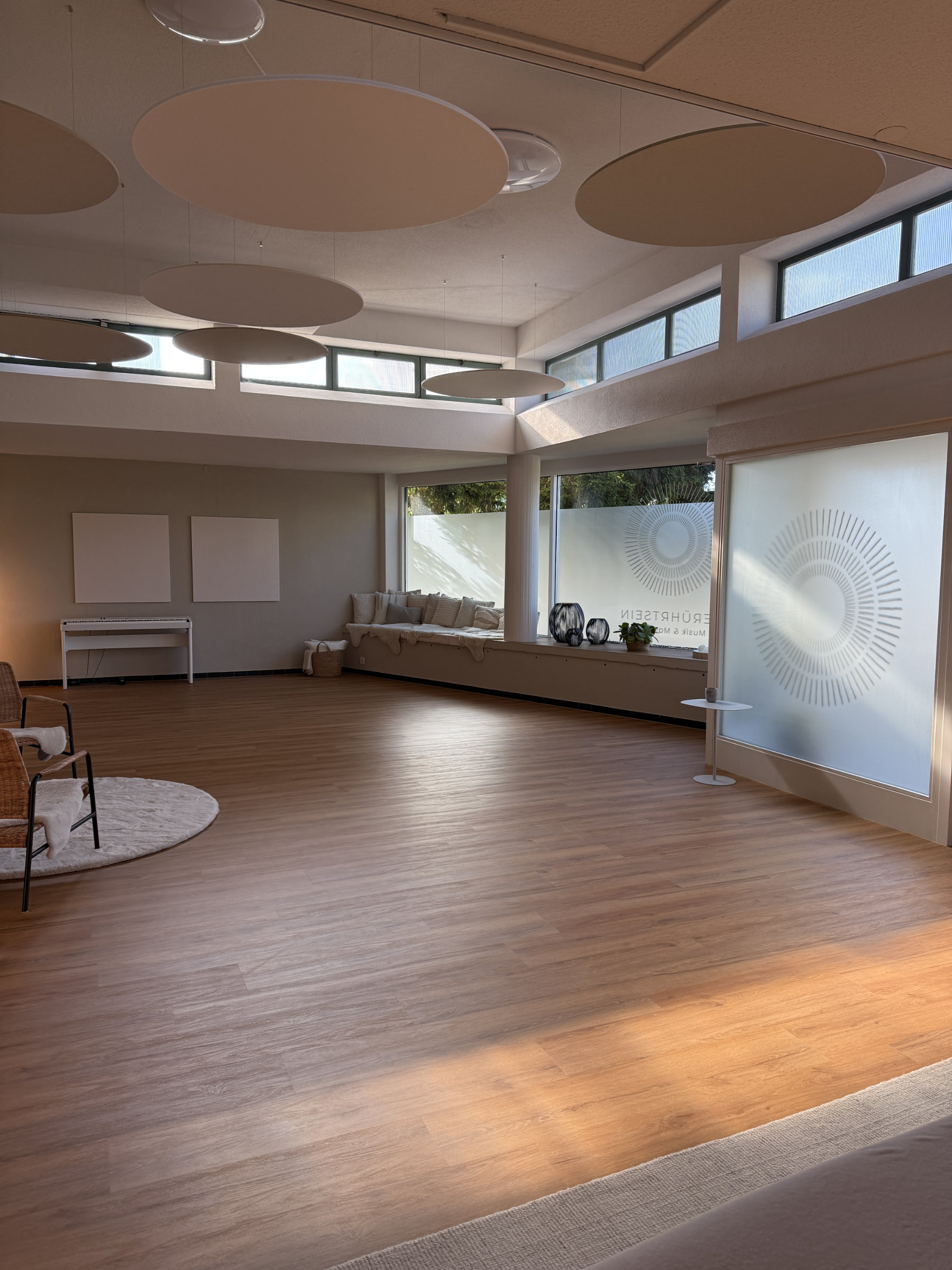 Der Raum Berührtsein. Ein großer heller Raum mit Holzboden, Sitzbank mit Kissen am Fenster und runden, an der Decke hängenden Akustikpaneelen.